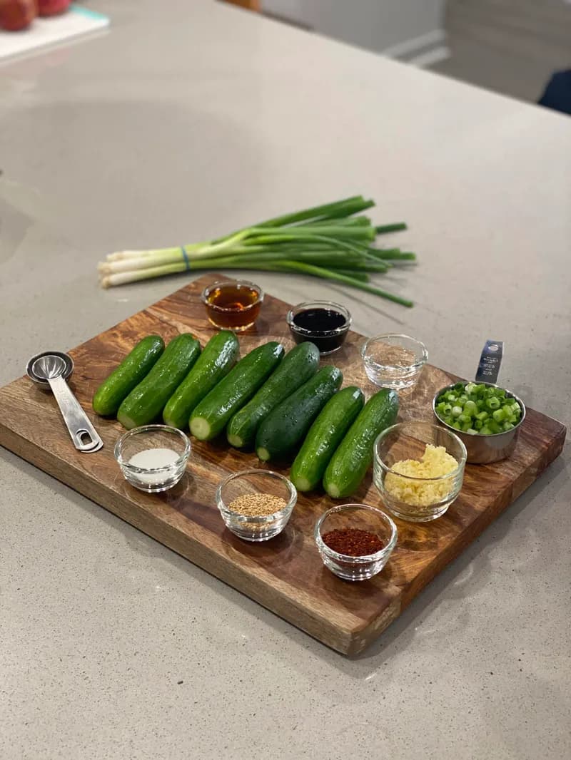 Mise en place for Korean cucumber salad — Persian cucumbers, gochugaru, sesame oil, soy sauce, garlic, scallions, and sesame seeds on a wooden cutting board