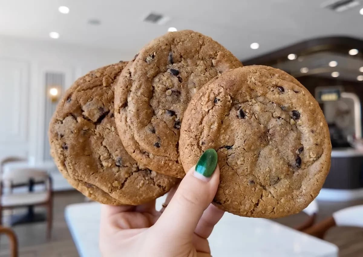 Hand holding three 85°C Bakery oatmeal chocolate chip cookies in front of the bakery interior
