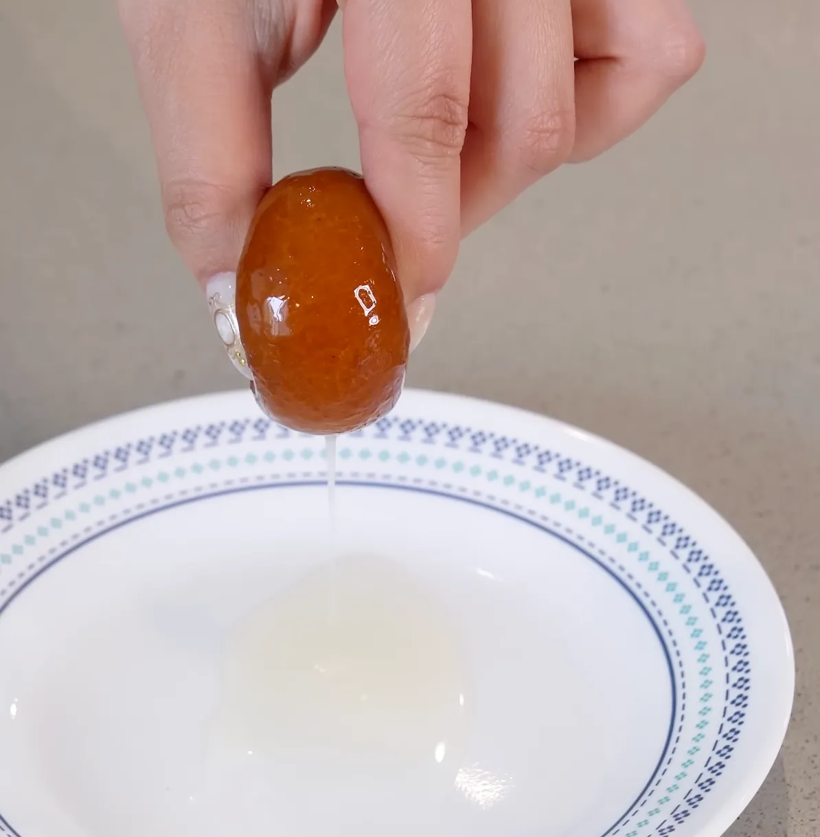 Hand holding a dark brown glazed gulab jamun ball over a white plate with syrup dripping off