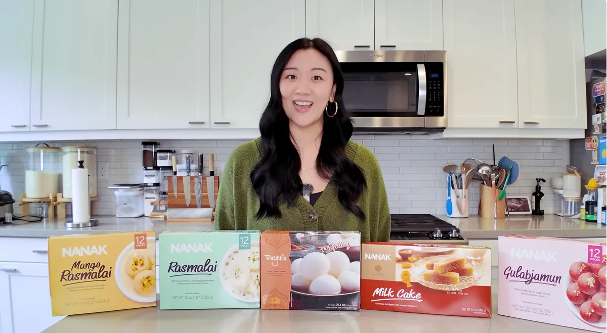 Jasmine standing in her kitchen behind all five Nanak Indian dessert boxes including mango rasmalai, rasmalai, rasgulla, milk cake, and gulab jamun