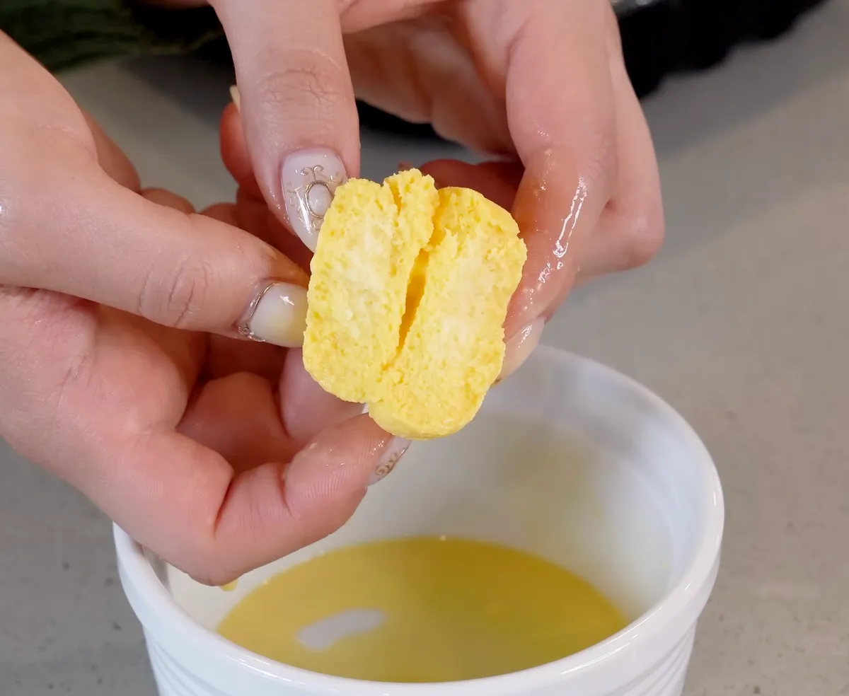 Hands pulling apart a mango rasmalai disc to reveal its yellow interior above a white cup