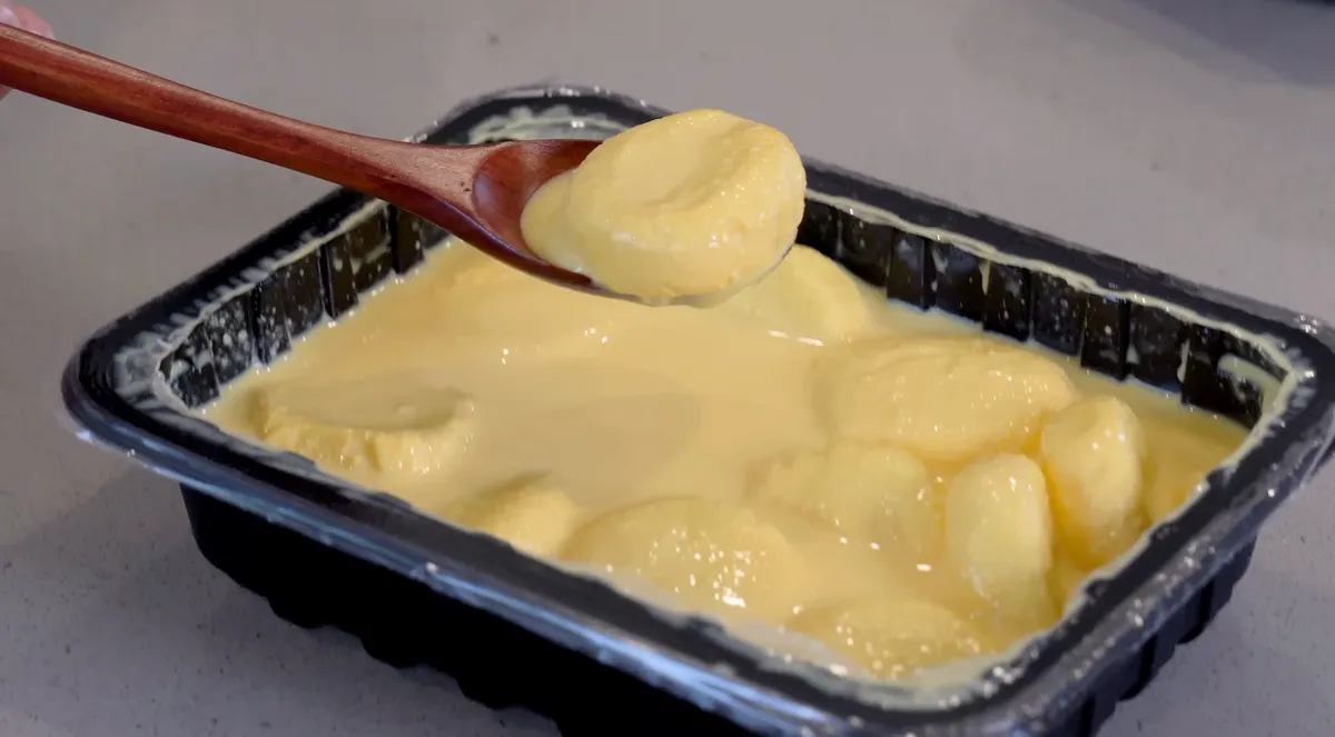 A mango rasmalai disc on a wooden spoon above a tray of rasmalai in yellow mango sauce