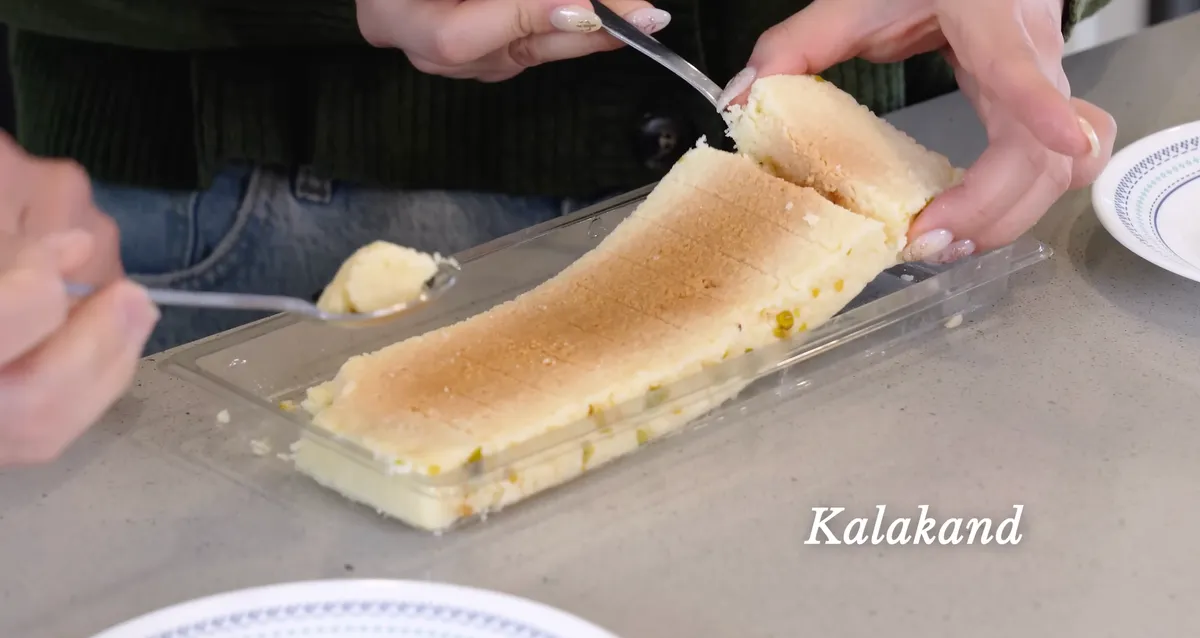 Two hands cutting into a block of Nanak milk cake kalakand with a fork, showing its dense crumbly texture