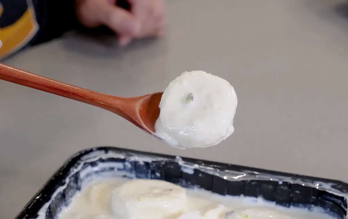 A single Nanak rasmalai disc balanced on a wooden spoon above a tray of rasmalai in creamy white sauce