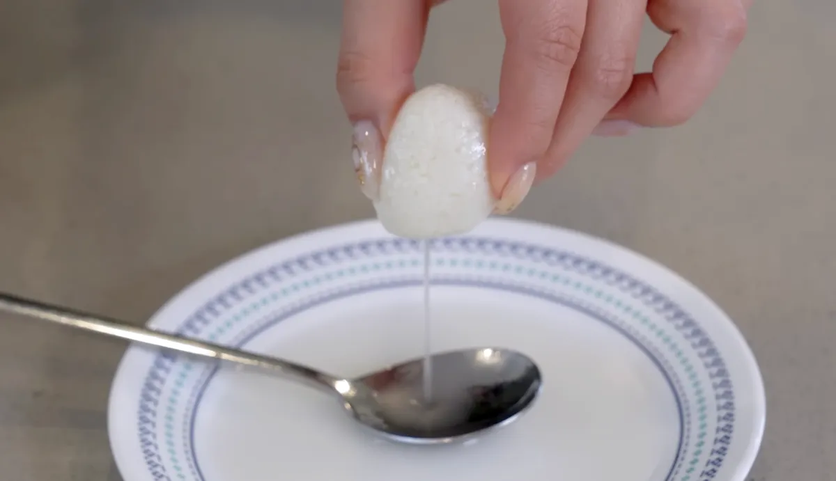 Hand squeezing a rasgulla ball over a white plate, with sugar syrup dripping down