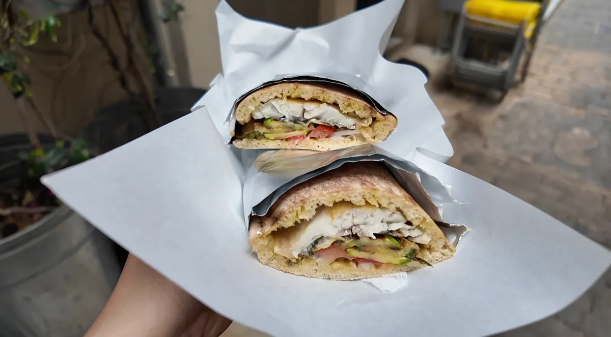 Atlantikos fish sandwich cut in half and wrapped in white paper, showing grilled fish, avocado, and tomato filling