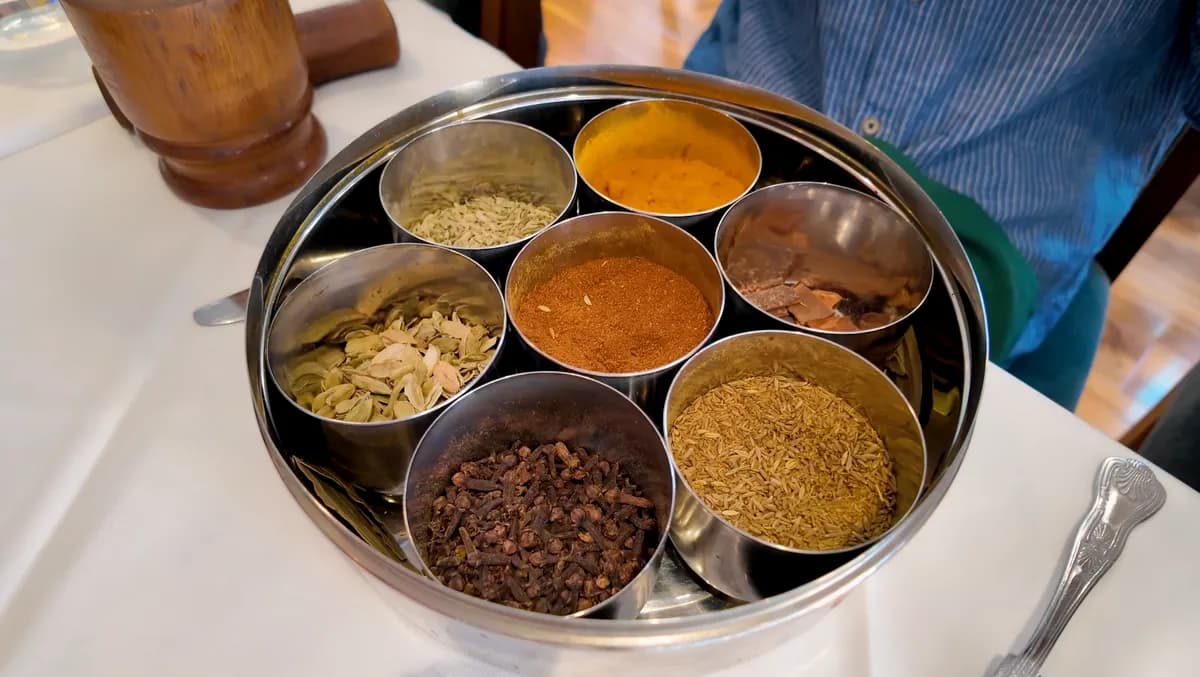 Steel spice tray with seven individual bowls of whole spices including cloves, cumin, cardamom, turmeric, and fennel used in the Brick Lane food tour spice guessing game